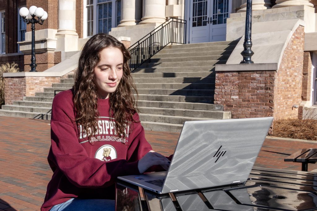 Girl with brown hair sits at an outdoor table in front of her laptop.