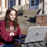 Girl with brown hair sits at an outdoor table in front of her laptop.